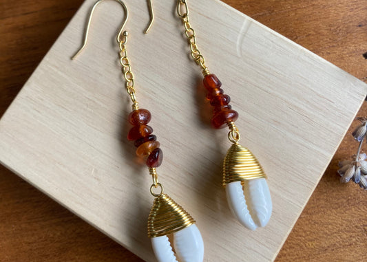 Hessonite Garnet and cowrie gold coloured copper wire findings, dangle drop earrings, displayed on wooden block with lavender flowers slightly in frame